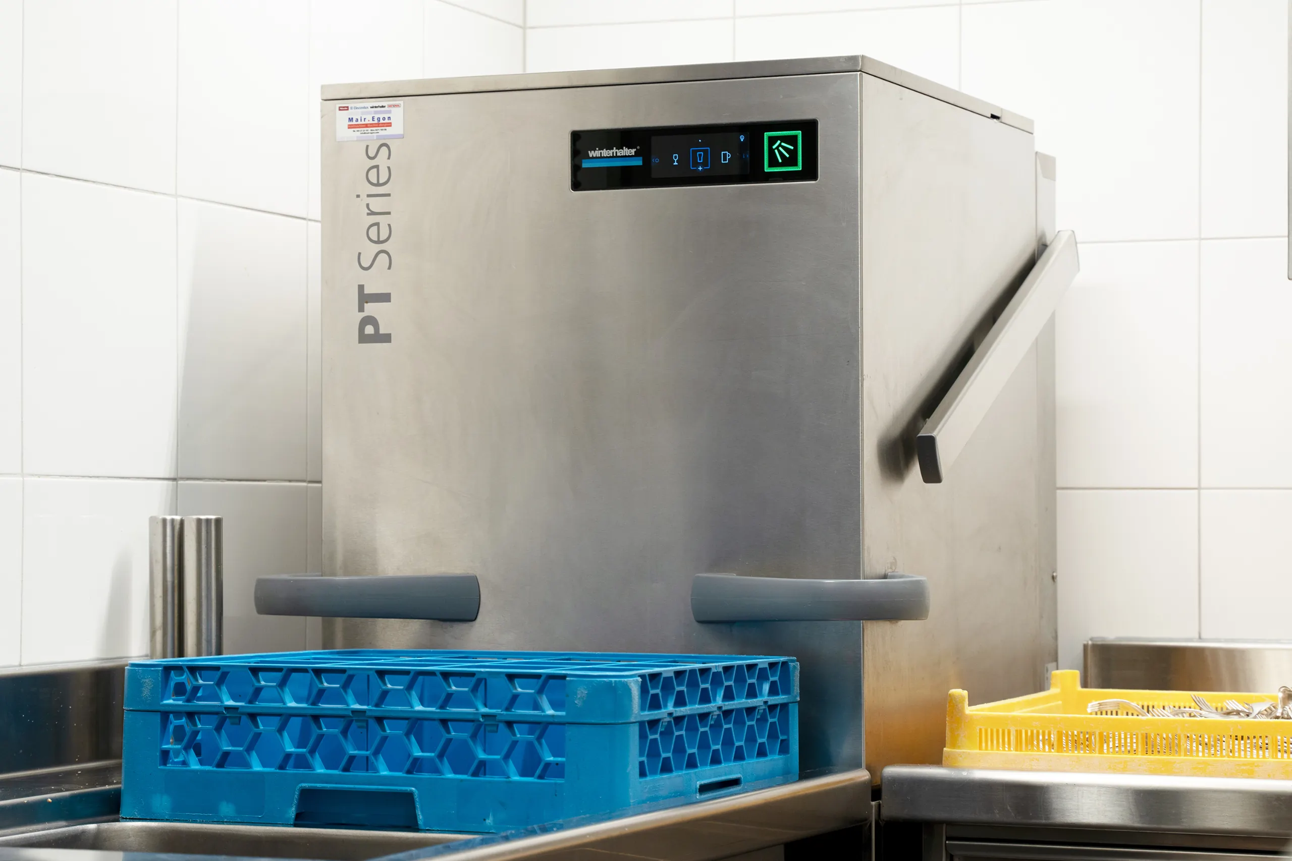 Eine Spülmaschine steht in der Ecke einer Küche. Links davon ist eine blaue Kiste und rechts davon eine gelbe Kiste mit Besteck.